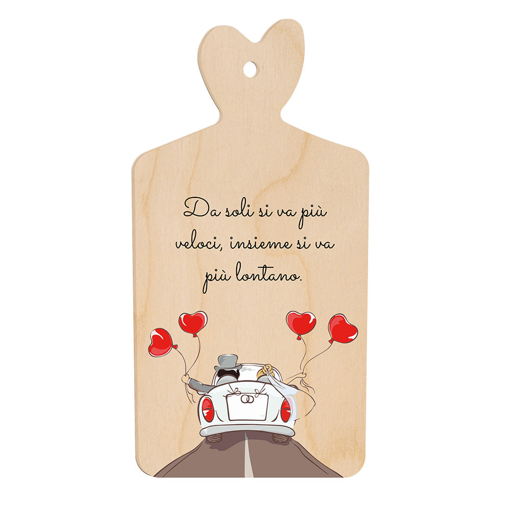 Tagliere in legno a forma di cuore con frase “Da soli si va più veloci, insieme si va più lontano” e illustrazione di sposi in auto con palloncini rossi a cuore.