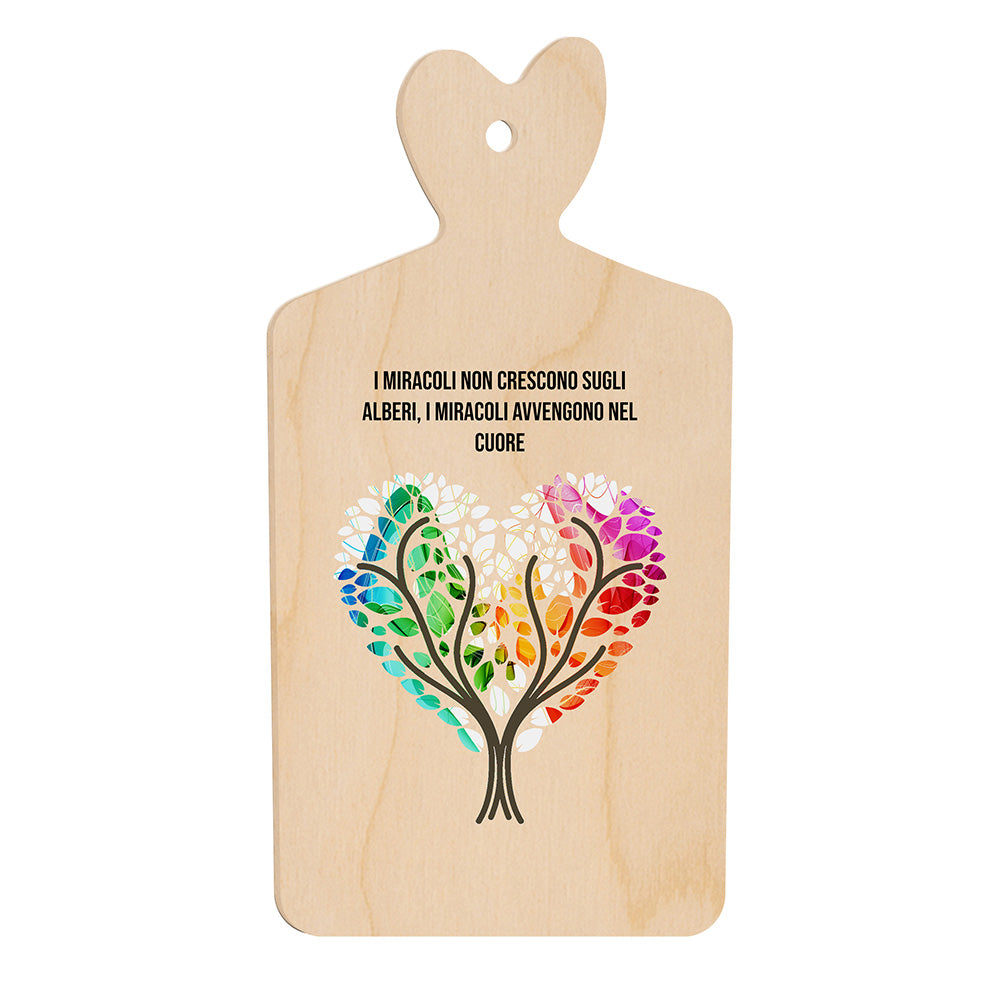 Tagliere in legno a forma di cuore con frase “I miracoli non crescono sugli alberi, i miracoli avvengono nel cuore” e illustrazione di due alberi che formano un cuore colorato.