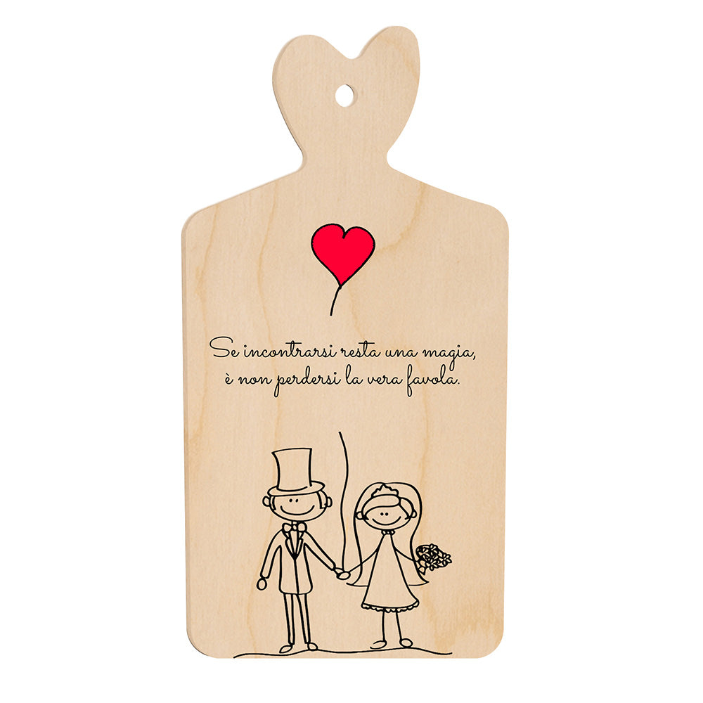 Tagliere in legno a forma di cuore con frase romantica e illustrazione stilizzata di sposi che si tengono per mano sotto un palloncino rosso a forma di cuore.