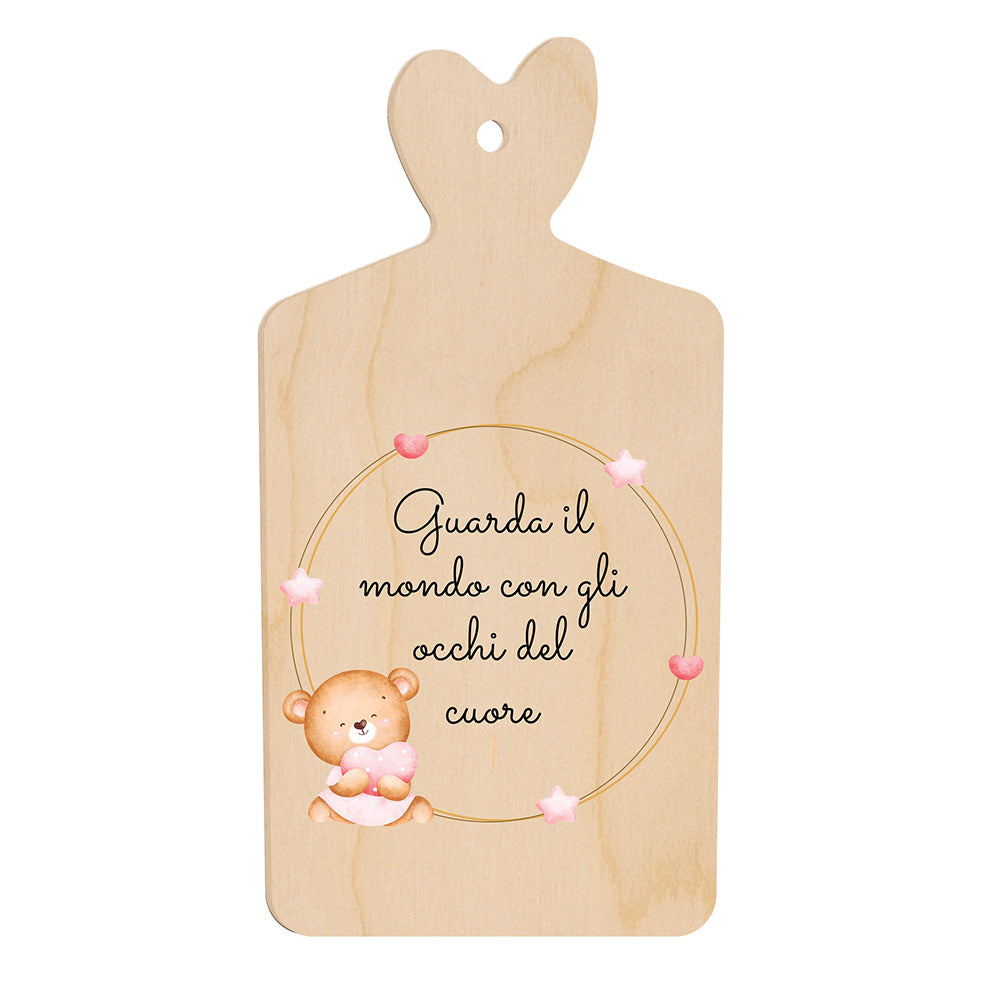 Tagliere in legno a forma di cuore con frase “Guarda il mondo con gli occhi del cuore” e illustrazione di un orsetto seduto accanto a una cornice di stelline rosa.
