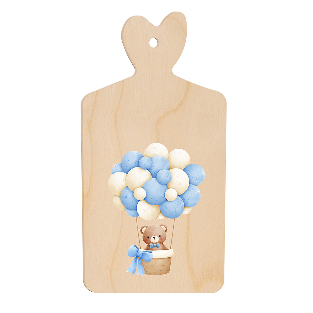Tagliere in legno a forma di cuore con illustrazione di un orsetto in mongolfiera fatta di palloncini azzurri e bianchi, in stile dolce e infantile.