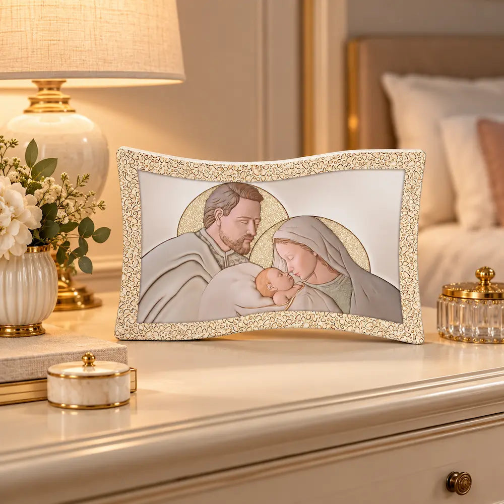 Bomboniera Sacra Famiglia in tonalità beige e oro, ambientata su comò elegante in camera da letto con lampada e fiori bianchi.