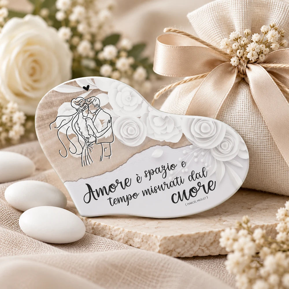 Bomboniera matrimonio a forma di cuore con coppia di sposi e frase sull’amore, ambientata con sacchetto, confetti e fiori bianchi.