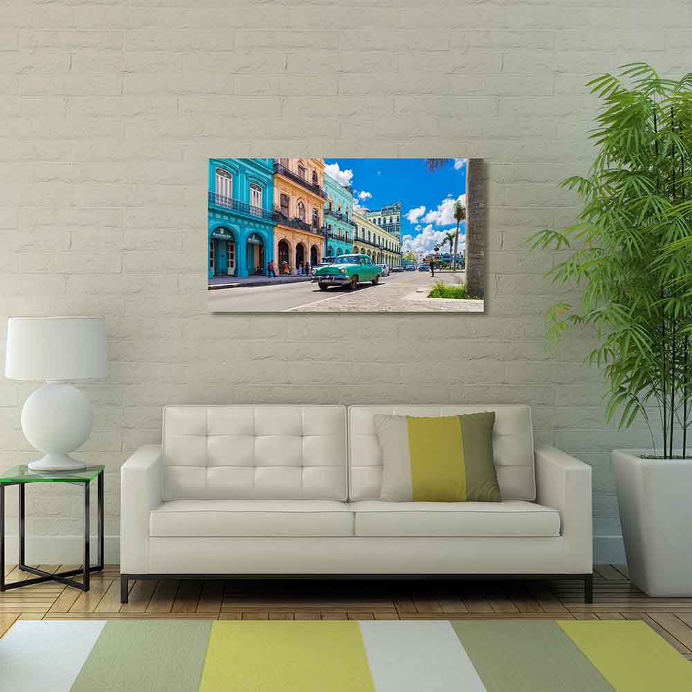 Quadro in plexiglass o vetro con auto vintage e palazzi dai colori accesi, appeso in soggiorno chiaro con divano moderno e pianta verde.