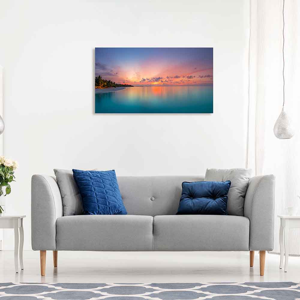 Quadro in plexiglass o vetro con paesaggio marino, cielo sfumato rosa-arancio e mare cristallino, appeso al muro sopra un divano.