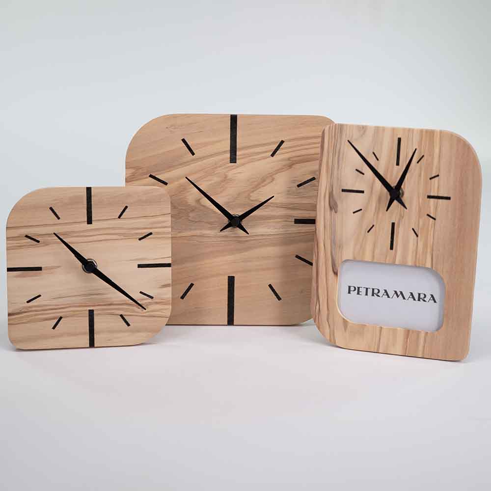 Tre orologi in legno d’ulivo con forme arrotondate, venature naturali ben visibili e indici neri su quadranti chiari dal design moderno ed essenziale.