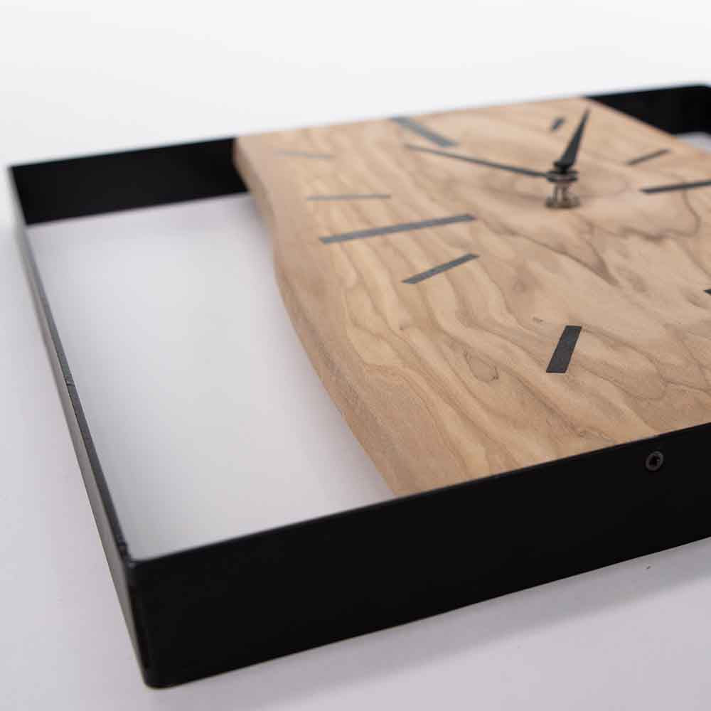 Orologio da parete in legno di ulivo e acciaio, visto da un angolo, evidenziando il contrasto tra i materiali naturali e moderni.