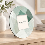 Portafoto rotondo in vetro con decoro geometrico verde Shapes, ambientato su una madia in legno chiaro dal design moderno