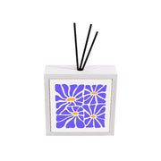 Profumatore ceramica bianco con piastrella floreale, quattro grandi margherite in blu pervinca e centri arancio su sfondo bianco, bastoncini reed neri