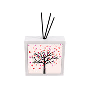 Profumatore ceramica bianco con albero dai rami neri spogli, cuori colorati in rosa rosso e viola come foglie cadenti, sfondo rosa chiaro, reed neri