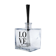 Profumatore ambiente vetro quadrato, etichetta minimal con scritta LOVE in caratteri bold neri e frase "is the answer" in corsivo con cuore, reed neri