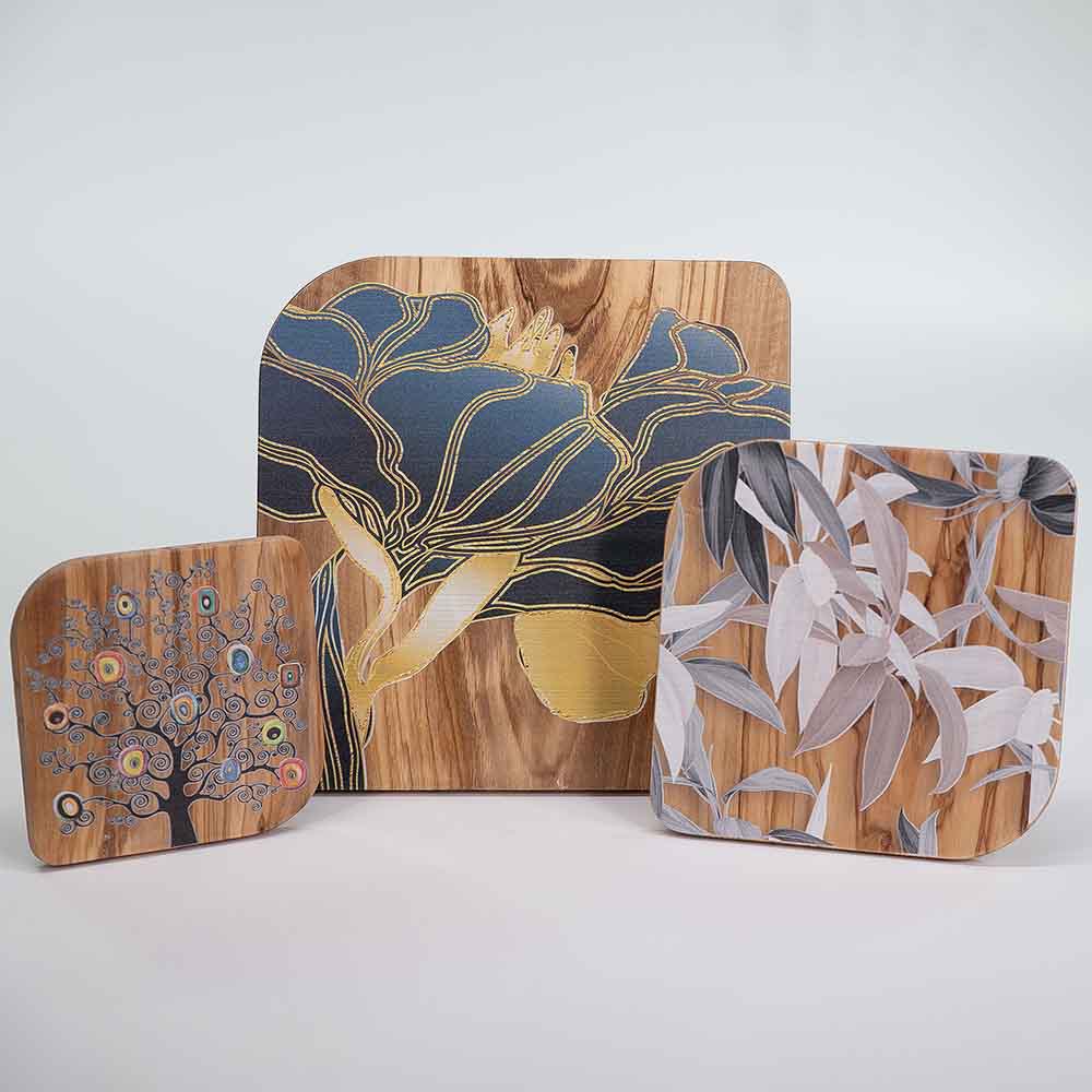 Tre quadretti in legno d'ulivo con decori: fiore blu e oro, albero stilizzato con cerchi colorati e foglie in tonalità neutre su sfondo naturale.
