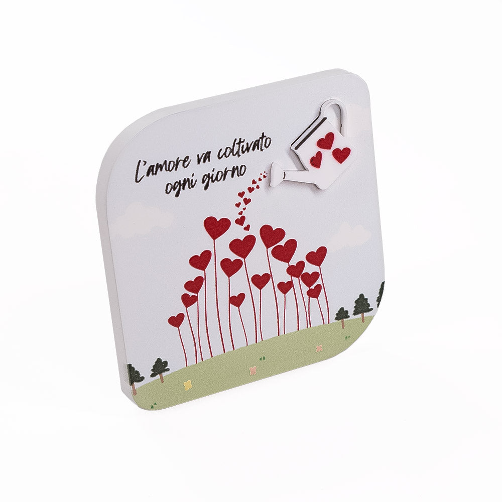 Decorazione in legno vista laterale, con base d'appoggio e rilievo evidente su annaffiatoio e cuori rossi, frase romantica sulla superficie.