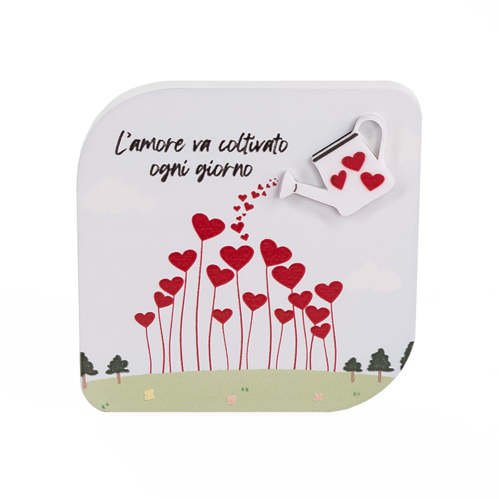 Quadretto in legno con annaffiatoio in rilievo che spruzza cuori rossi su steli a forma di fiori, con scritta "L’amore va coltivato ogni giorno".