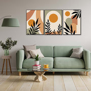 Tris di quadri appesi in fila sopra un divano verde: forme beige/terracotta e foglie nere con sole ocra.