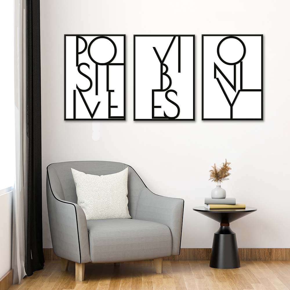 Tris di quadri appesi in fila sopra una poltrona grigia: lettering nero “POSITIVE”, “VIBES” e “ONLY” su bianco.