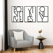Tris di quadri appesi in fila sopra una poltrona grigia: lettering nero “POSITIVE”, “VIBES” e “ONLY” su bianco.