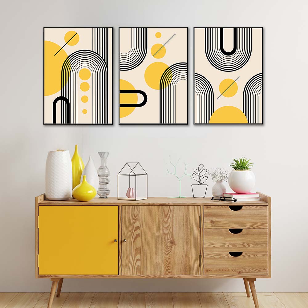 Tris di quadri appesi in fila sopra una madia: archi a righe nere e cerchi gialli su fondo beige, mood retrò.