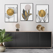 Tris di quadri appesi in fila sopra una madia: foglie e fiori a linea nera con cerchi oro e un cerchio beige rosato.