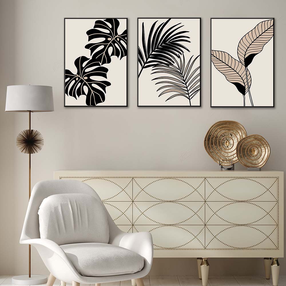 Tris di quadri appesi in fila sopra una credenza: foglie tropicali nere e beige (monstera e palma) su fondo chiaro.