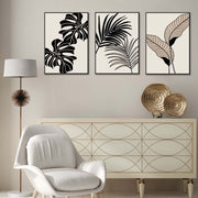 Tris di quadri appesi in fila sopra una credenza: foglie tropicali nere e beige (monstera e palma) su fondo chiaro.