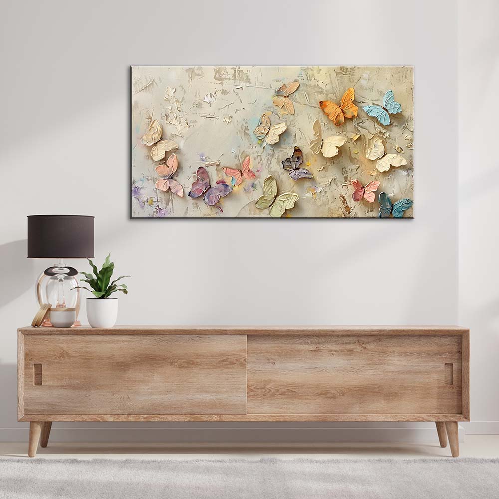 Quadro materico con farfalle a rilievo dai colori pastello, appeso su parete chiara sopra mobile in legno, con lampada e pianta in soggiorno.