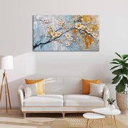 Quadro materico con fiori bianchi a rilievo e tocchi oro su sfondo azzurro e ocra, appeso sopra divano chiaro in soggiorno con cuscini gialli.