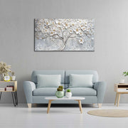 Quadro materico con albero fiorito bianco appeso centrato sopra divano azzurro in soggiorno moderno, fondo grigio con dettagli oro.