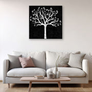 Quadro bianco e nero con albero stilizzato appeso a una parete chiara sopra un divano moderno beige, con cuscini decorativi e tavolino in legno.