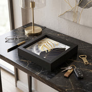 Svuotatasche in legno nero con foglie tropicali oro, argento e blu su fondo bianco, su consolle in marmo nero con occhiali e chiavi