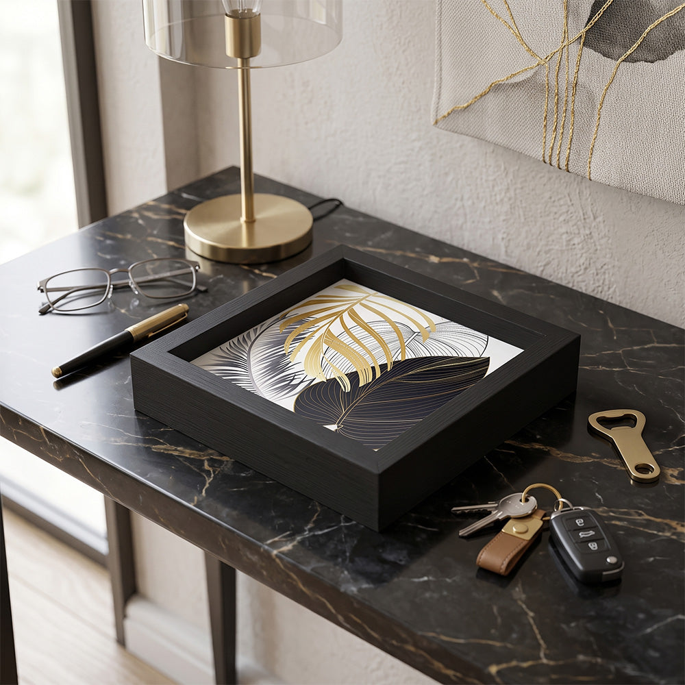 Svuotatasche in legno nero con foglie tropicali oro, argento e blu su fondo bianco, su consolle in marmo nero con occhiali e chiavi
