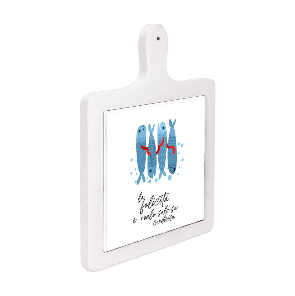 Tagliere verticale in ceramica bianca visto lateralmente, con pesci azzurri e frase “La felicità è reale solo se condivisa”.