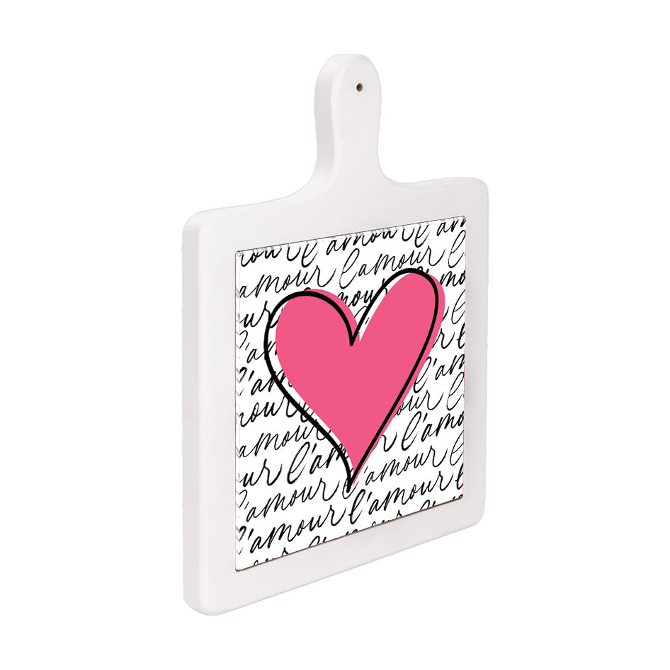 Tagliere verticale in ceramica bianca visto lateralmente, con grande cuore rosa e sfondo con scritta “l’amour” ripetuta in corsivo.