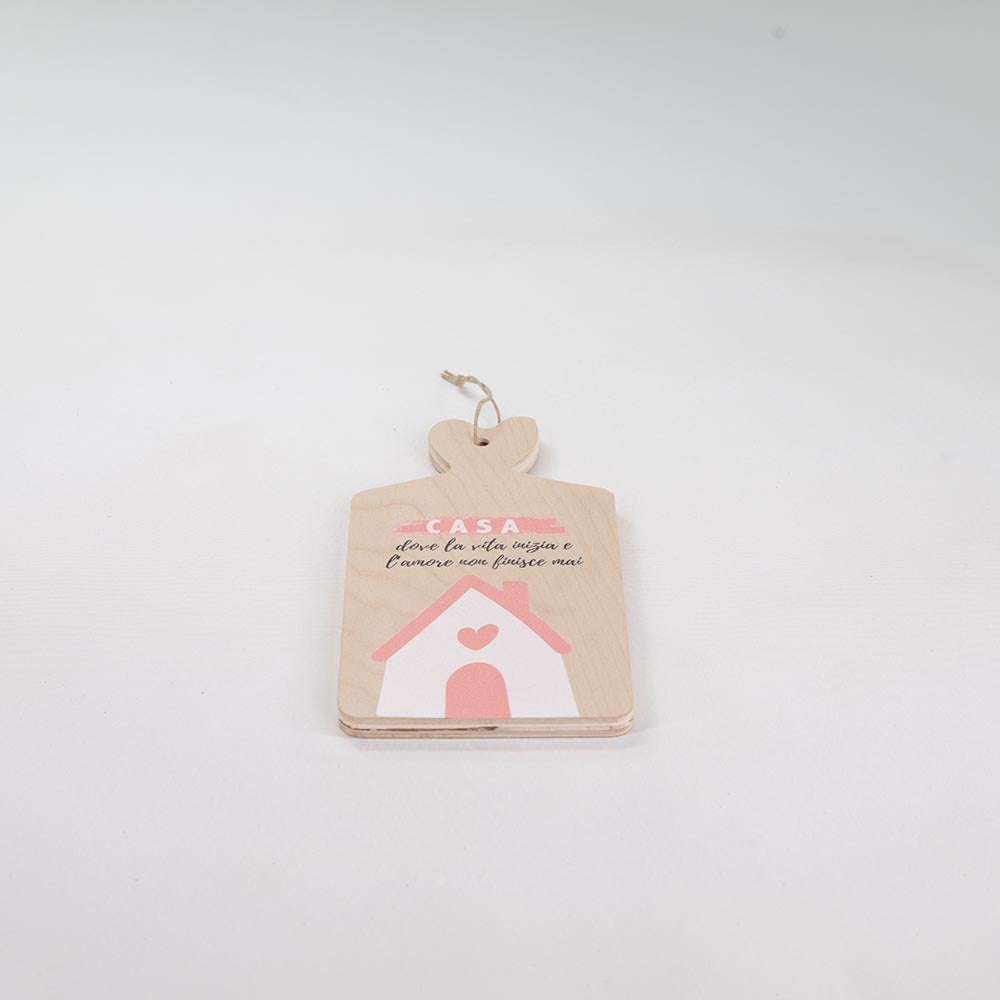 Tagliere in legno chiaro con manico a cuore, decorato con casetta rosa e frase sulla casa, posizionato disteso su sfondo bianco.