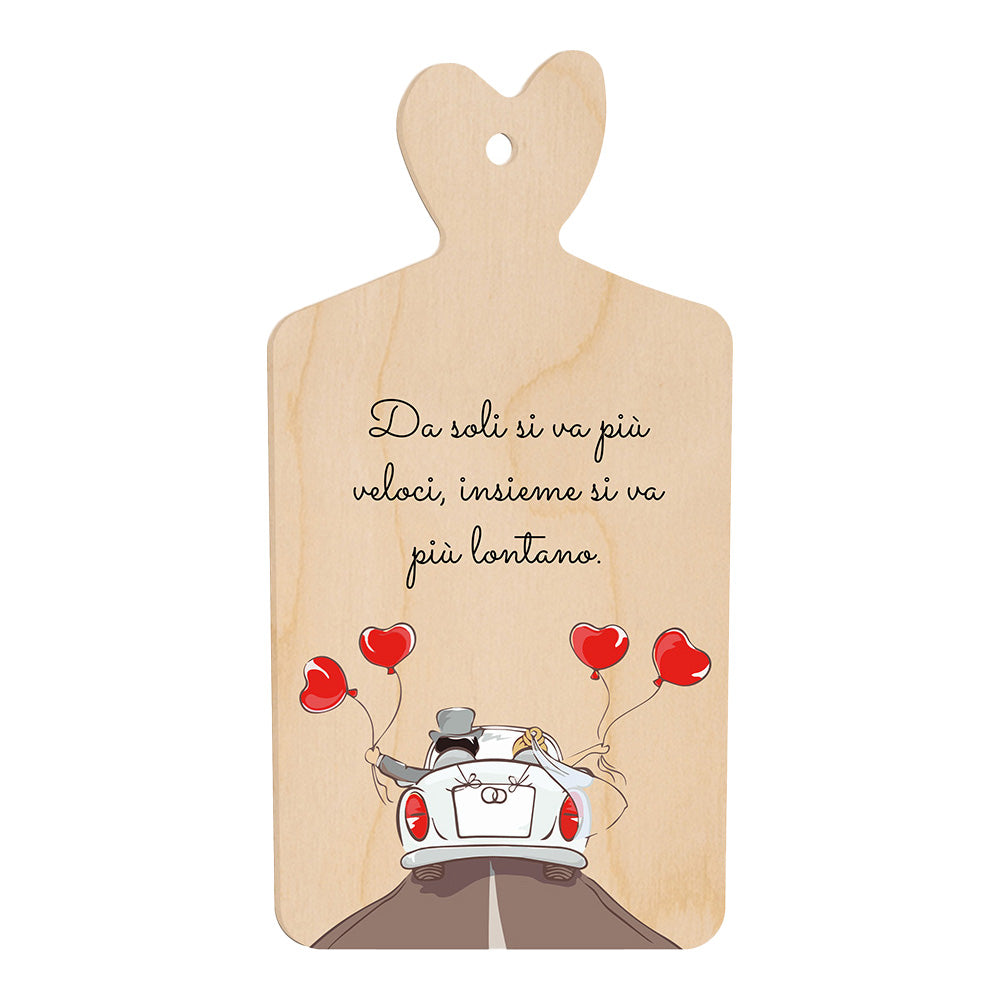 Tagliere in legno a forma di cuore con frase “Da soli si va più veloci, insieme si va più lontano” e illustrazione di sposi in auto con palloncini rossi a cuore.