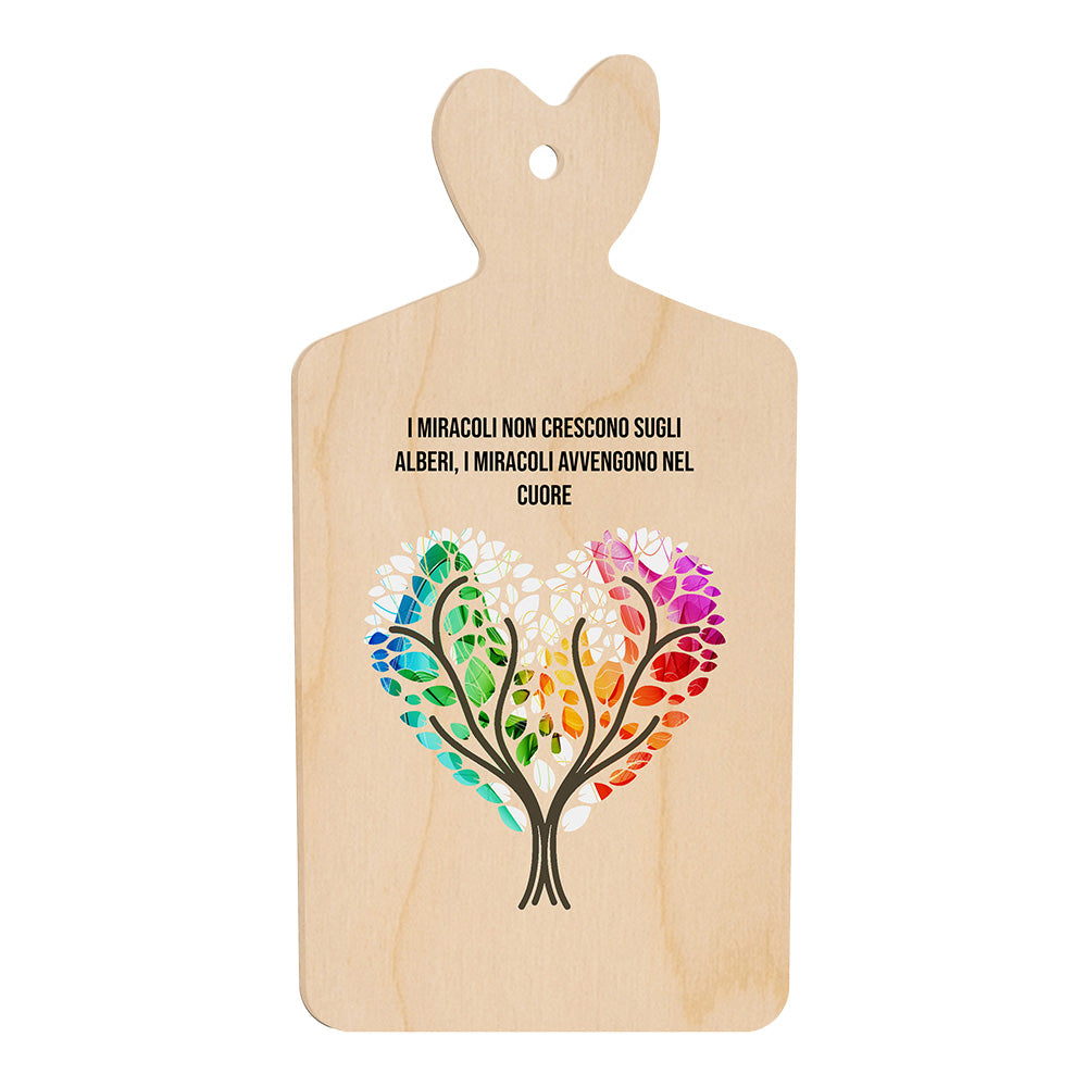 Tagliere in legno a forma di cuore con frase “I miracoli non crescono sugli alberi, i miracoli avvengono nel cuore” e illustrazione di due alberi che formano un cuore colorato.