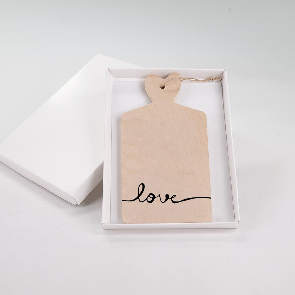 Tagliere decorativo in legno con manico a cuore e scritta "love" in corsivo nero, presentato in scatola regalo bianca.