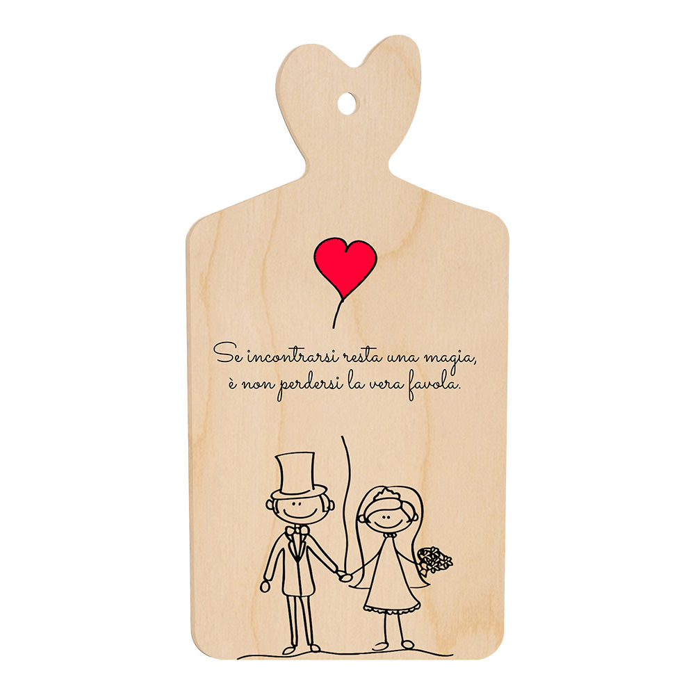 Tagliere in legno a forma di cuore con frase romantica e illustrazione stilizzata di sposi che si tengono per mano sotto un palloncino rosso a forma di cuore.
