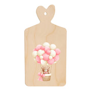 Tagliere in legno a forma di cuore con illustrazione di un orsetto in mongolfiera fatta di palloncini rosa e bianchi, in stile dolce e infantile.