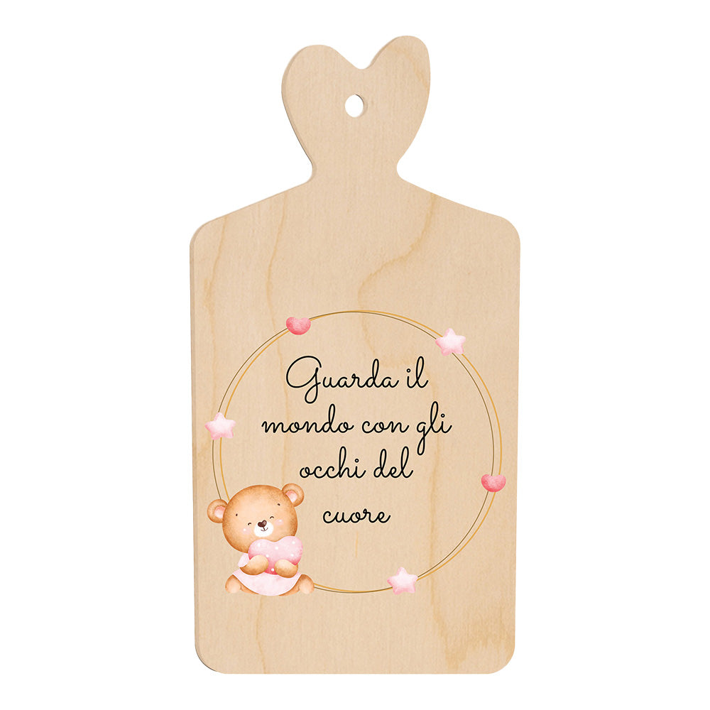 Tagliere in legno a forma di cuore con frase “Guarda il mondo con gli occhi del cuore” e illustrazione di un orsetto seduto accanto a una cornice di stelline rosa.